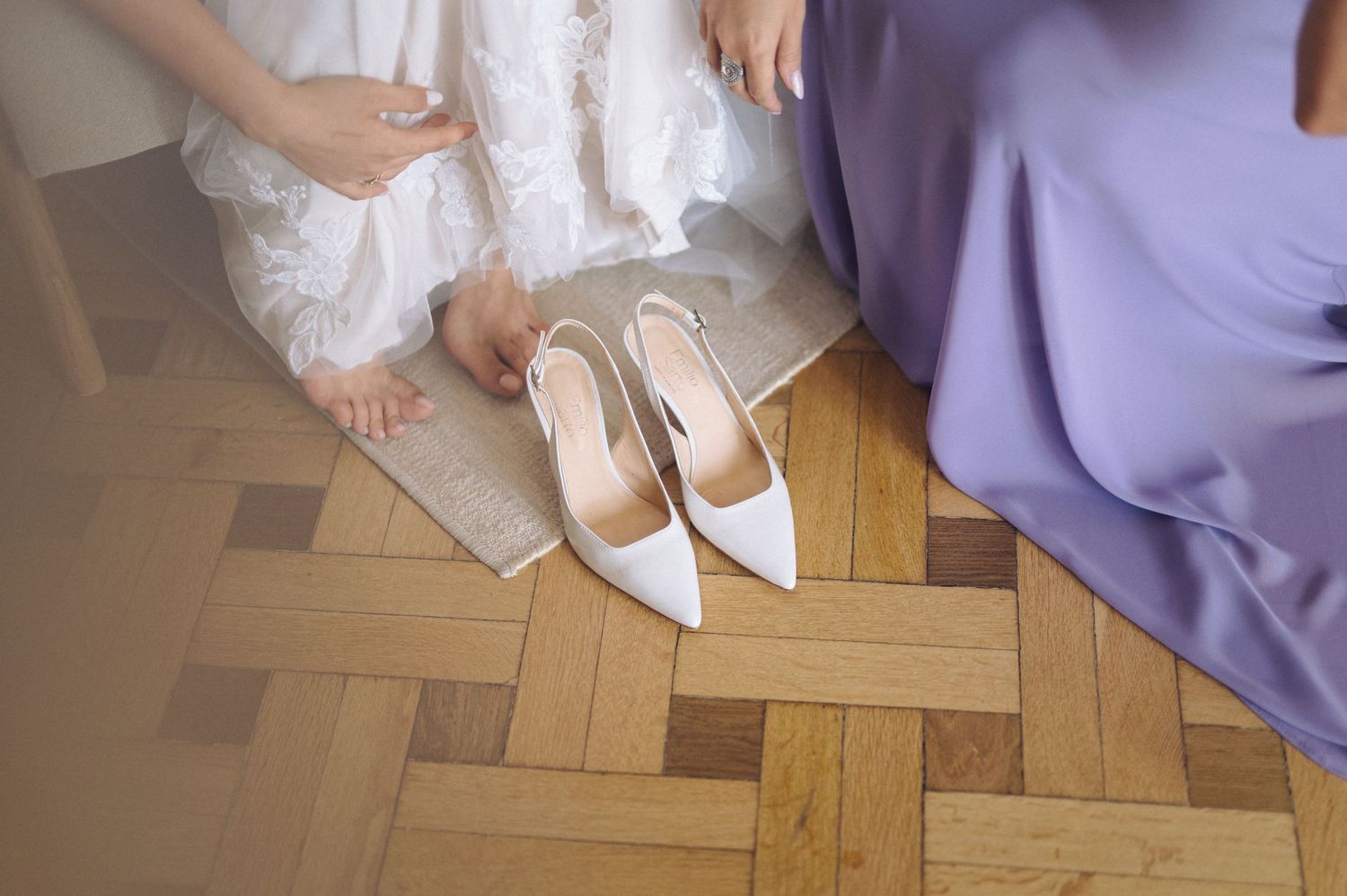 A bride in white shoes walks across a parquet wood floor with long purple bridesmaid dresses visible beside her.