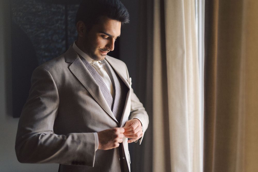 A person in a stylish gray suit adjusts their tie while standing near a bright window.