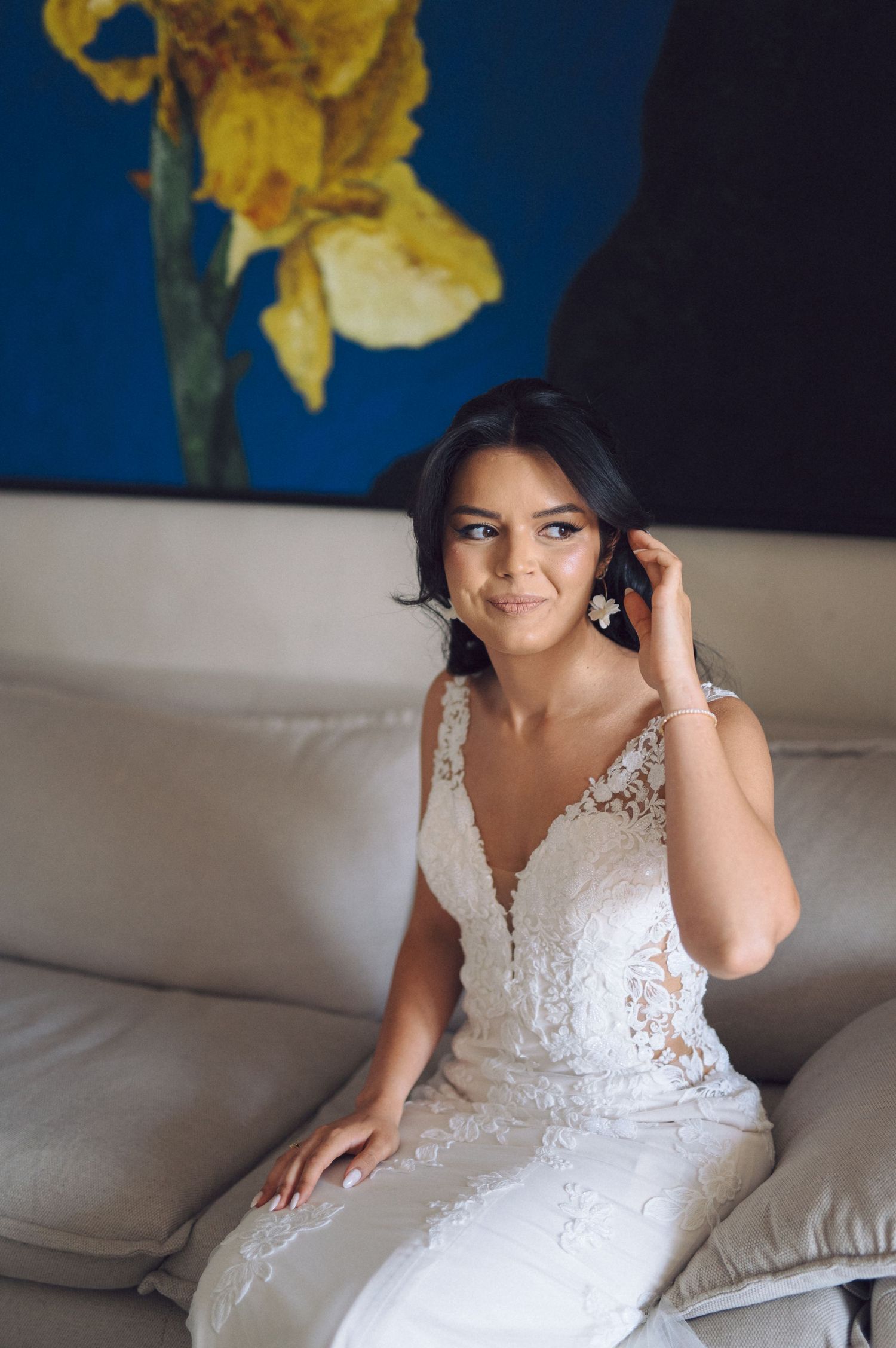 A bride in an elegant white lace wedding dress smiles while sitting on a gray couch with a yellow flower painting behind her.