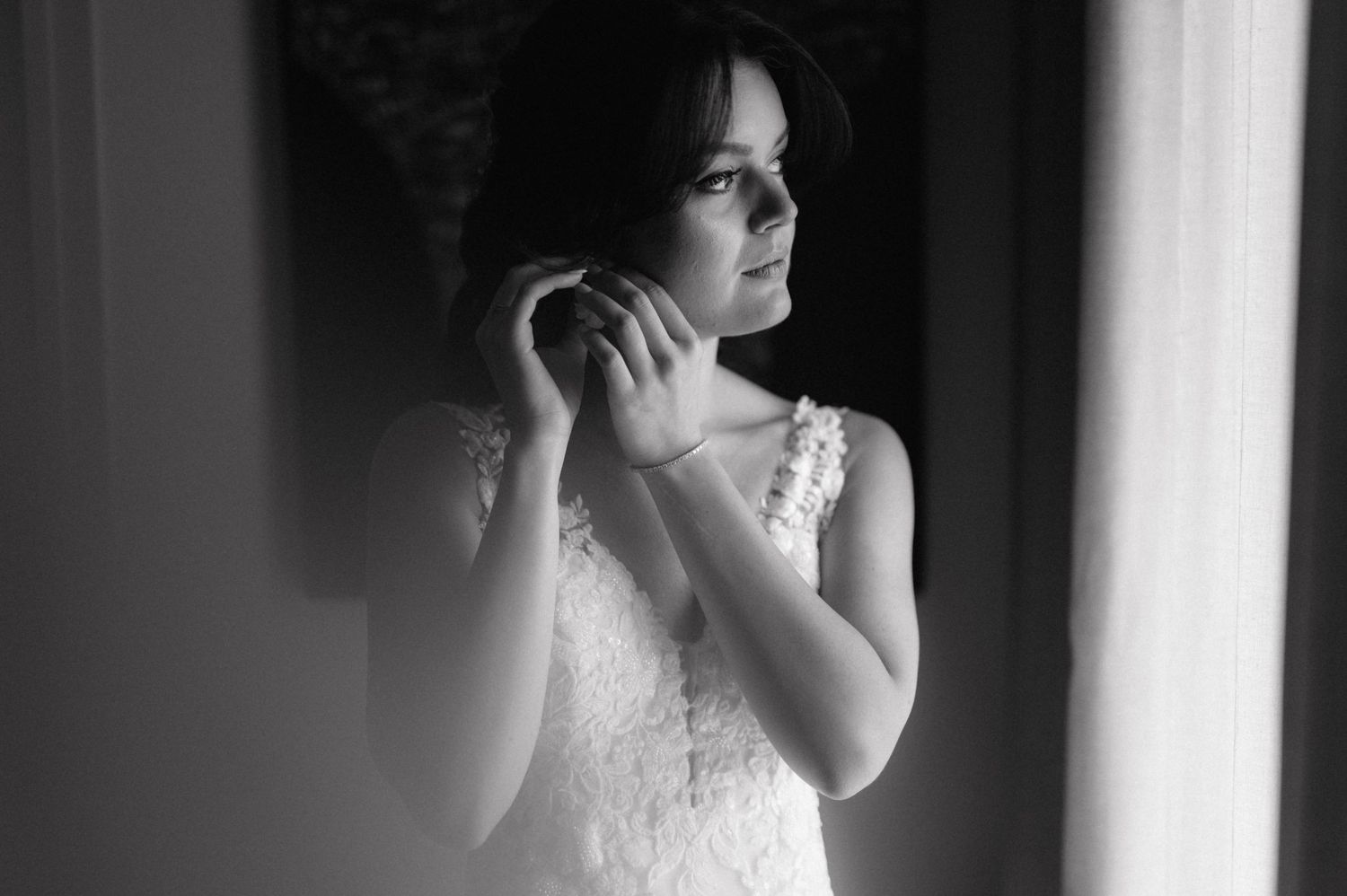 A bride in lace wedding dress stands by window light in dramatic black and white portrait.