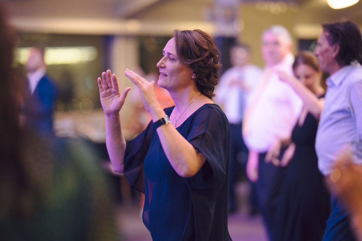 A person in a black top stands with raised hands in a prayer-like gesture at an indoor event.