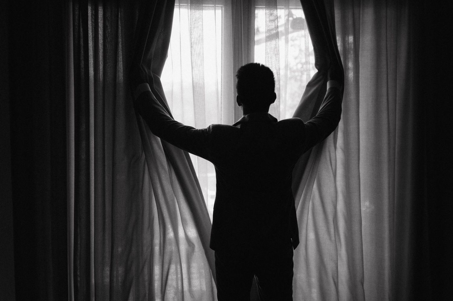 A silhouetted figure opens curtains by a bright window in a dark room creating a dramatic contrast.
