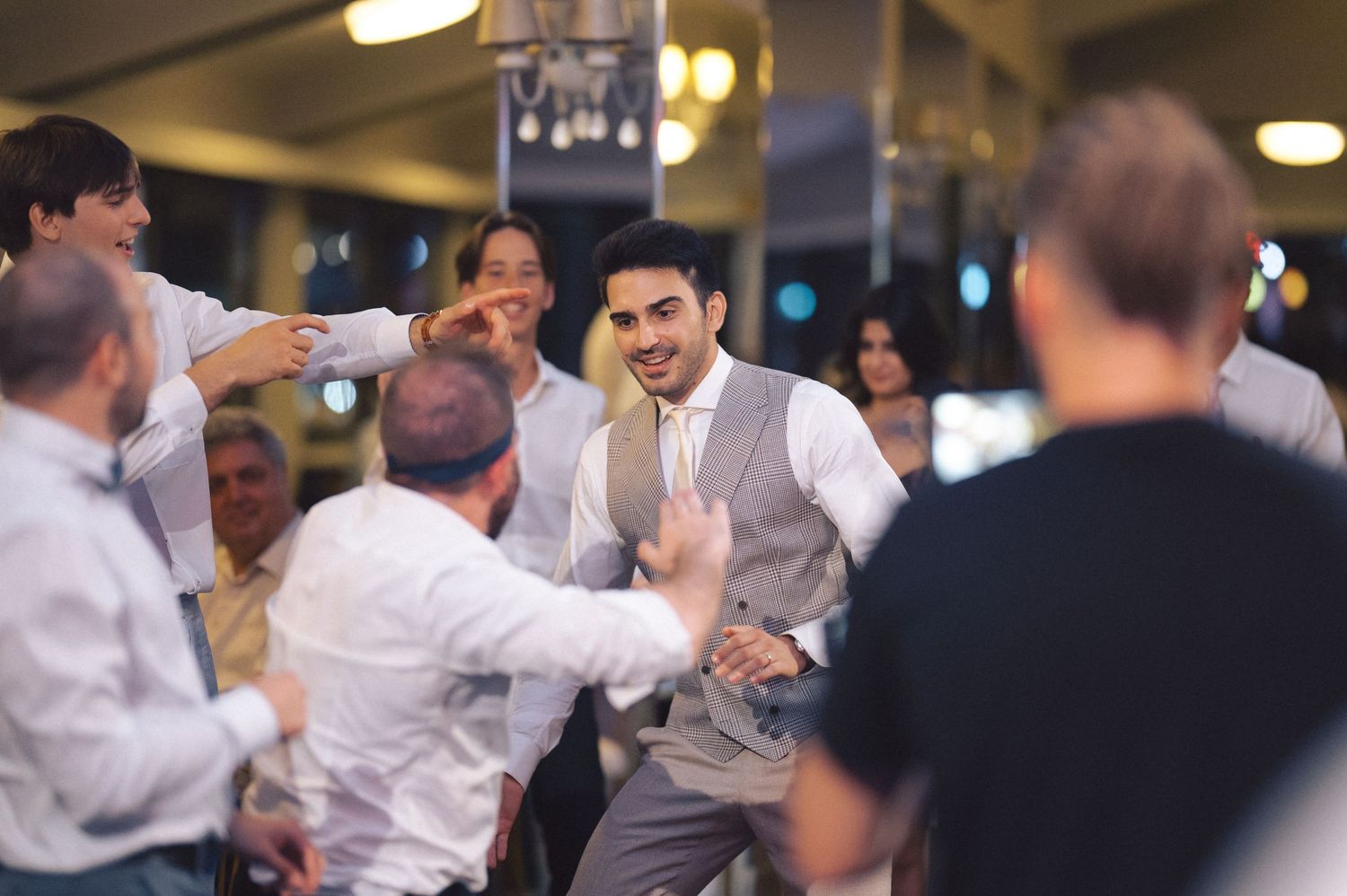 Wedding guests dance and celebrate enthusiastically at an indoor reception venue with warm lighting.