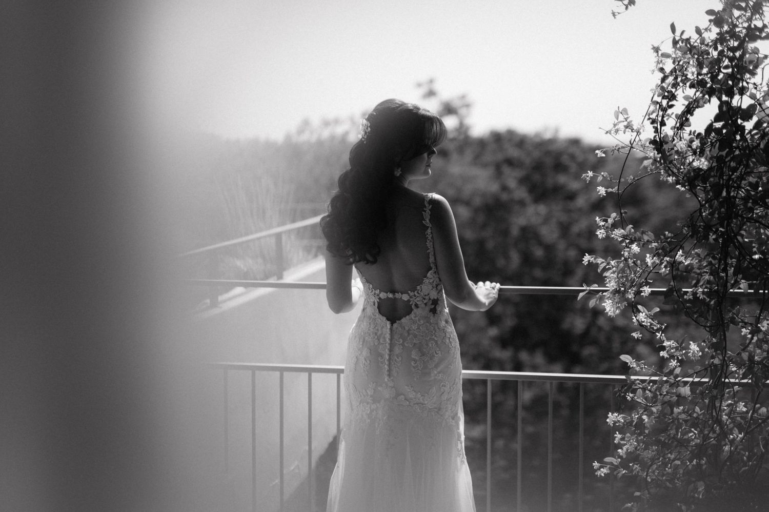 A figure in a flowing white dress stands backlit on a balcony in a dreamy black and white photograph.