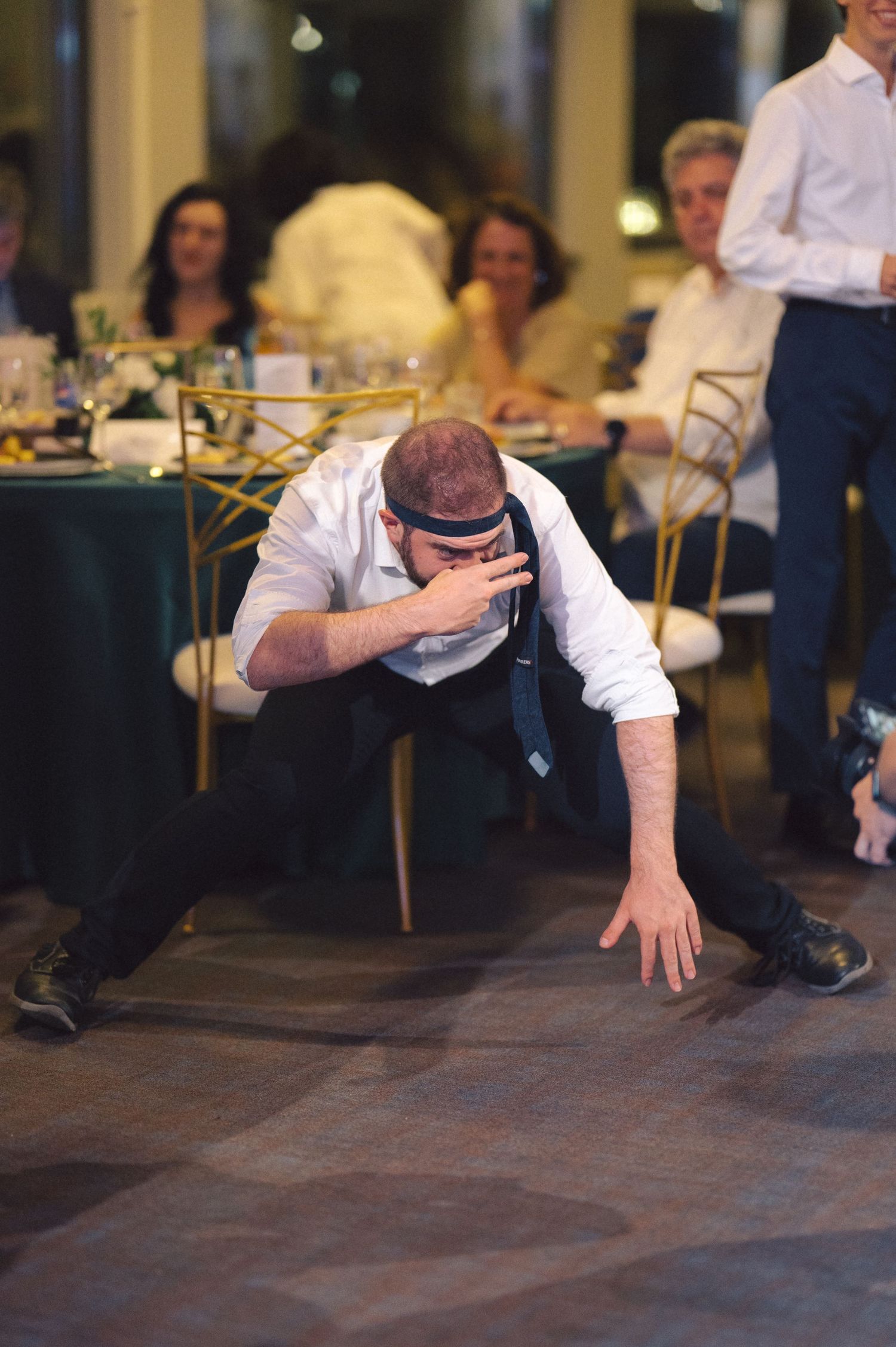 Person in white shirt and tie performs dramatic dance move in reception venue with guests seated at tables in background.