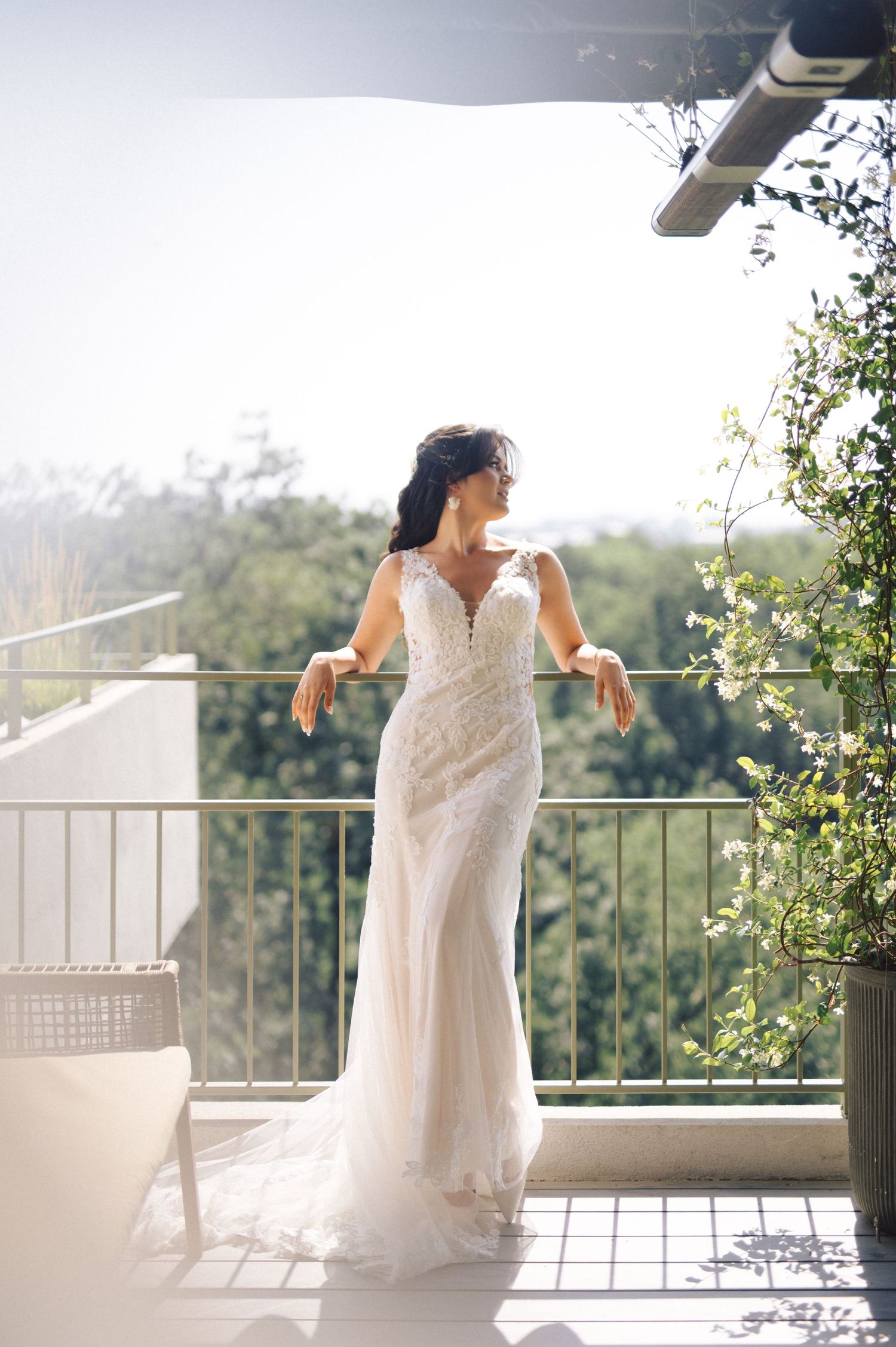 A bride in a fitted lace wedding gown stands elegantly on a sunny balcony overlooking a scenic view.