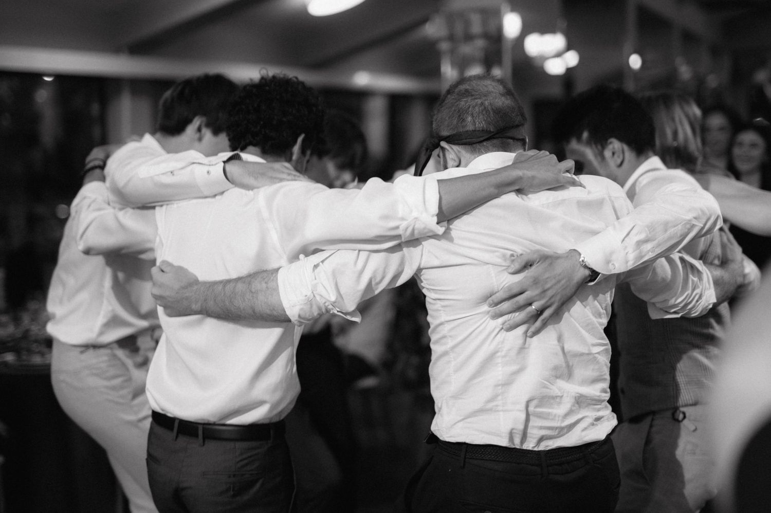 Black and white photo of groomsmen huddled together in a group embrace at a wedding reception.