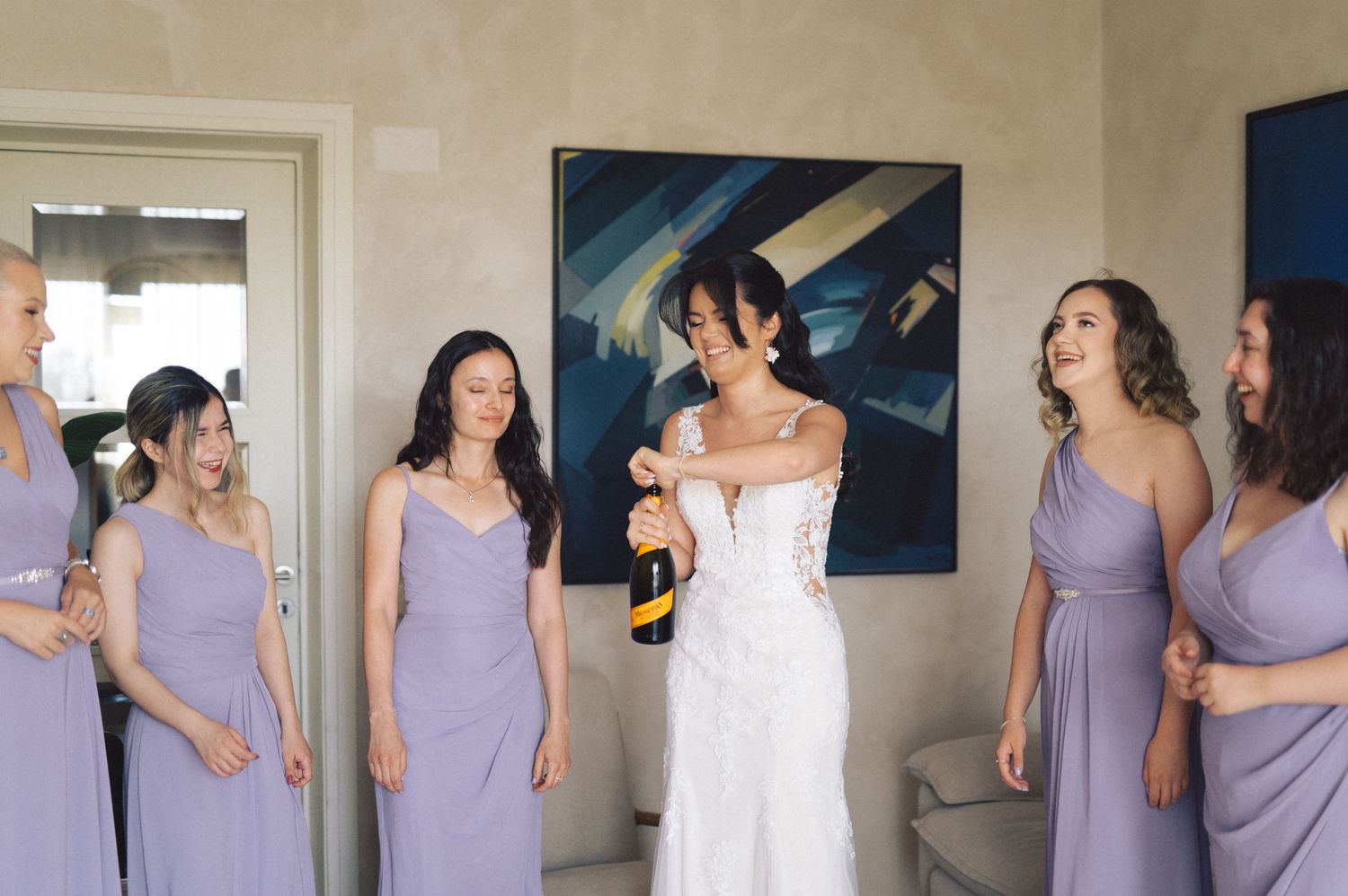 Bridesmaids in lavender dresses gather around a bride in a white gown during pre-wedding preparations.