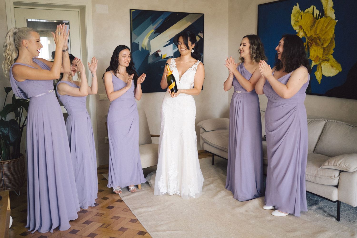 Several people in light purple bridesmaid dresses share joyful moments with someone wearing a white wedding gown.