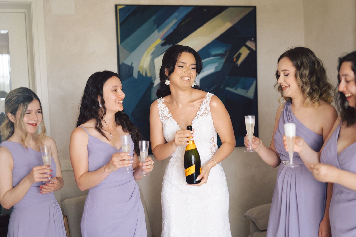 A group of women in lavender bridesmaid dresses share drinks and laughter with a bride in a white wedding gown.