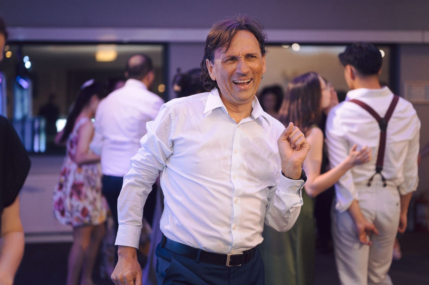 A group of people in white shirts dance together at an indoor social event with purple lighting.