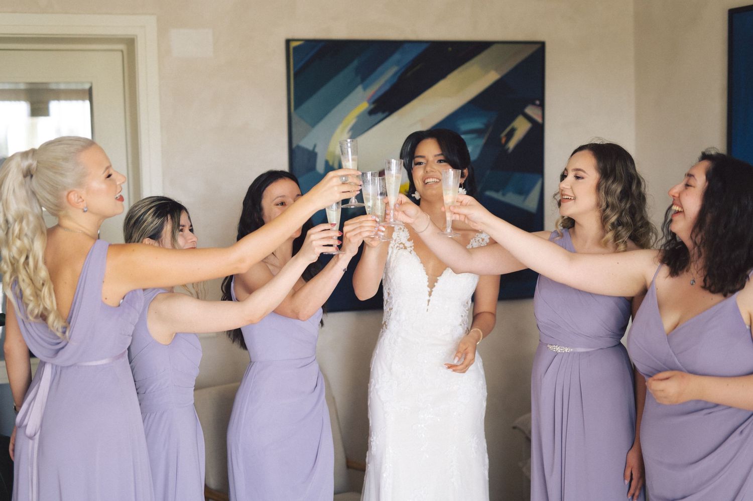 Bridesmaids in lavender dresses share celebratory moments getting ready before a wedding ceremony.