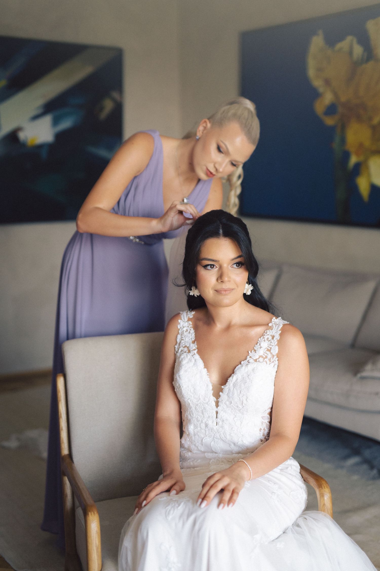 A bride in a white lace wedding dress gets her hair styled in an elegant preparation room setting.
