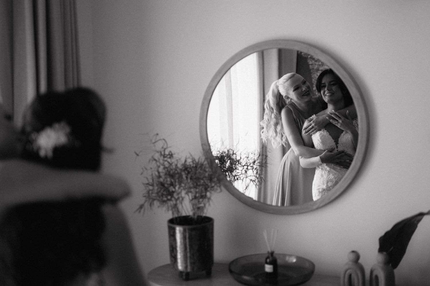 A tender embrace is reflected in a round mirror mounted on a wall beside a vase with flowers in black and white.
