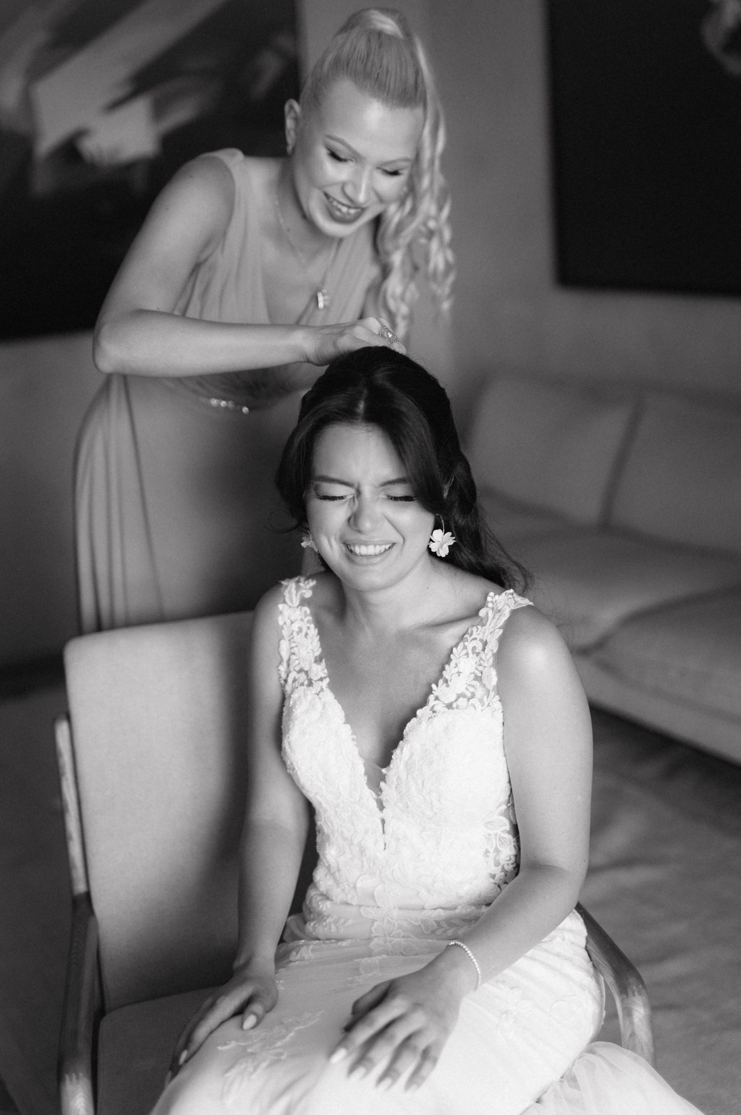 A black and white wedding photo captures a joyful moment as a bridesmaid helps style the bride's hair before the ceremony.