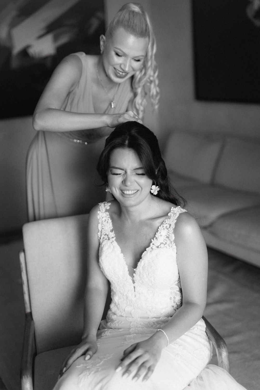 A black and white wedding photo captures a joyful moment as a bridesmaid helps style the bride's hair before the ceremony.