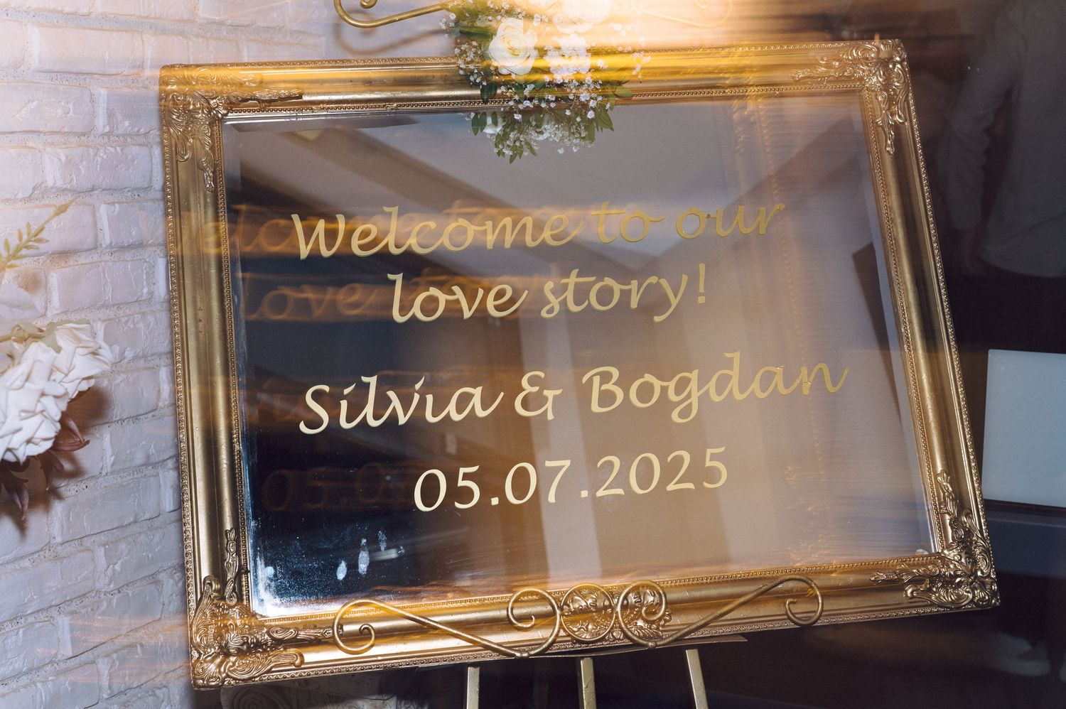 Elegant gold-framed welcome sign displaying names and wedding date on transparent glass with romantic lighting effect.