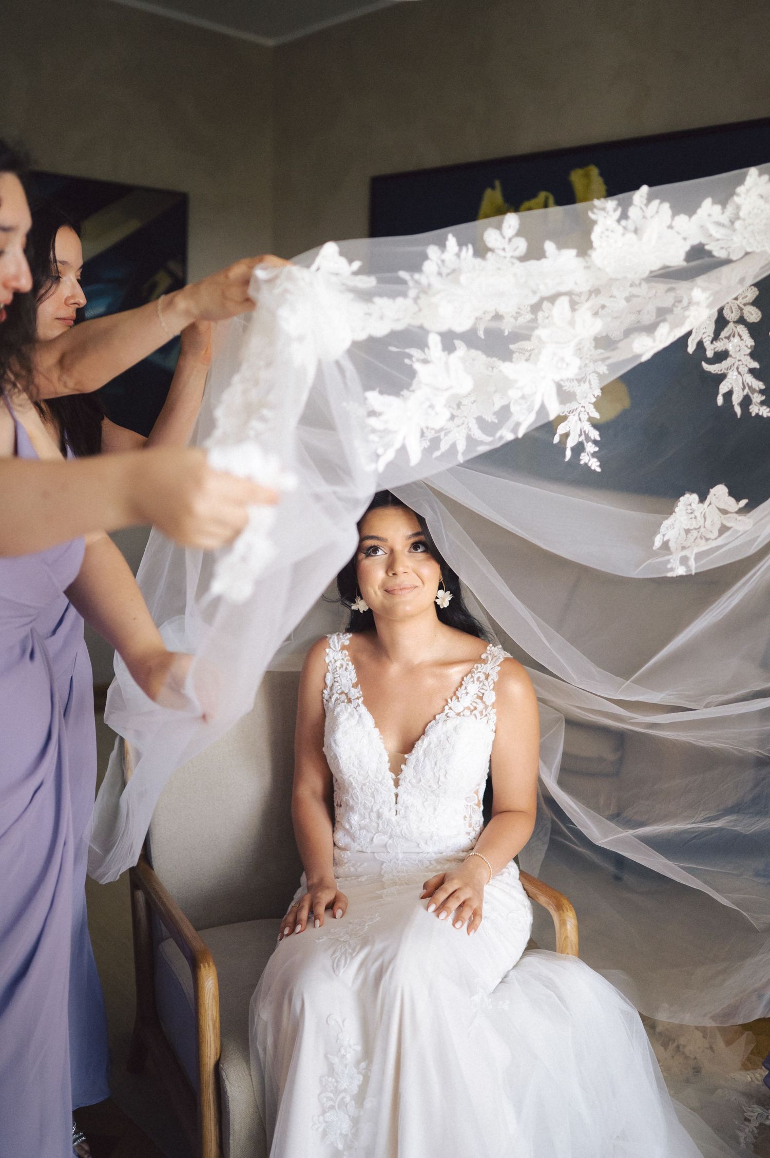 A bride in an elegant white lace wedding gown stands as assistants adjust her flowing veil.