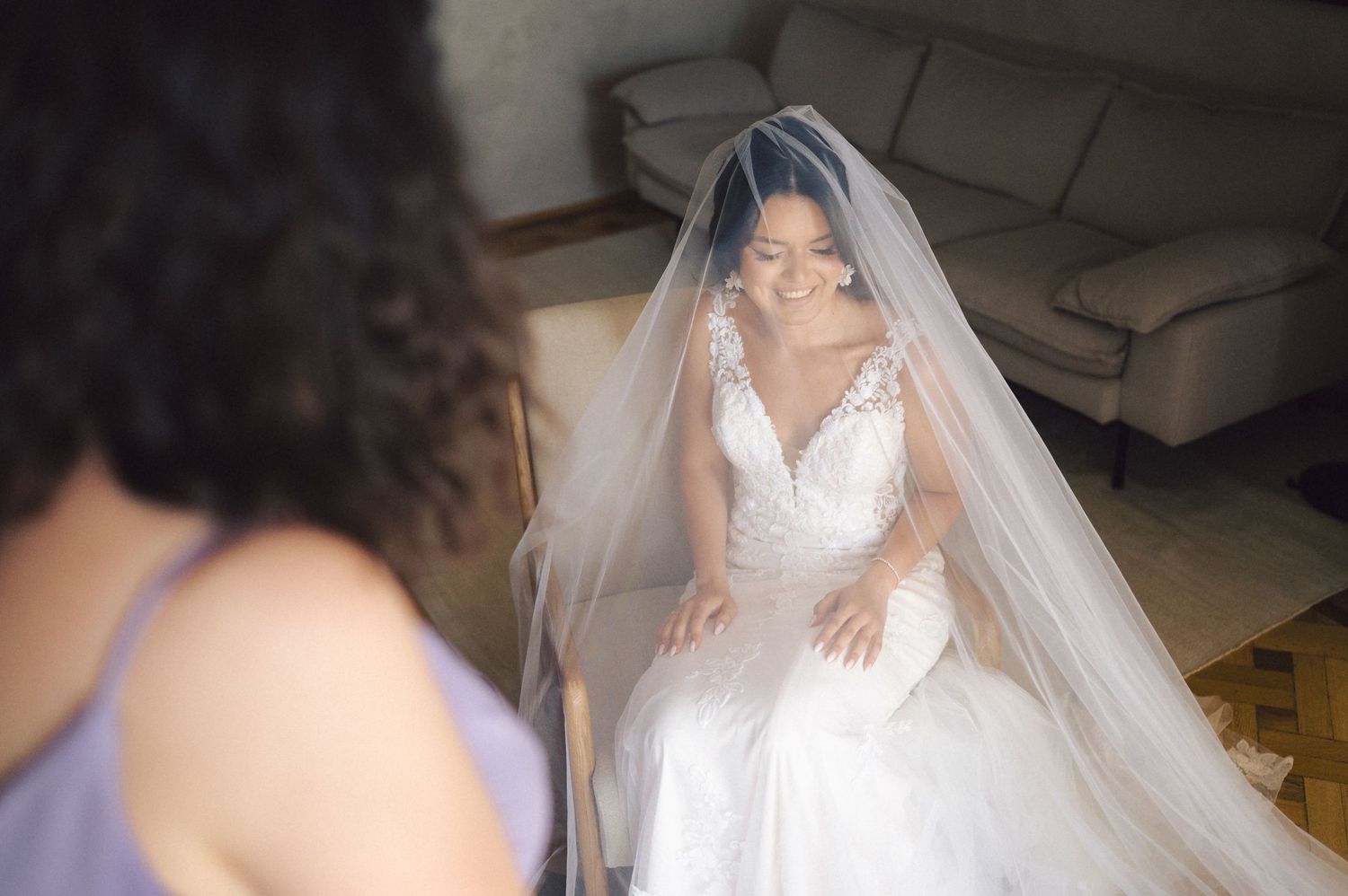 A bride in a white lace wedding gown with a flowing long veil sits and smiles during wedding preparations.