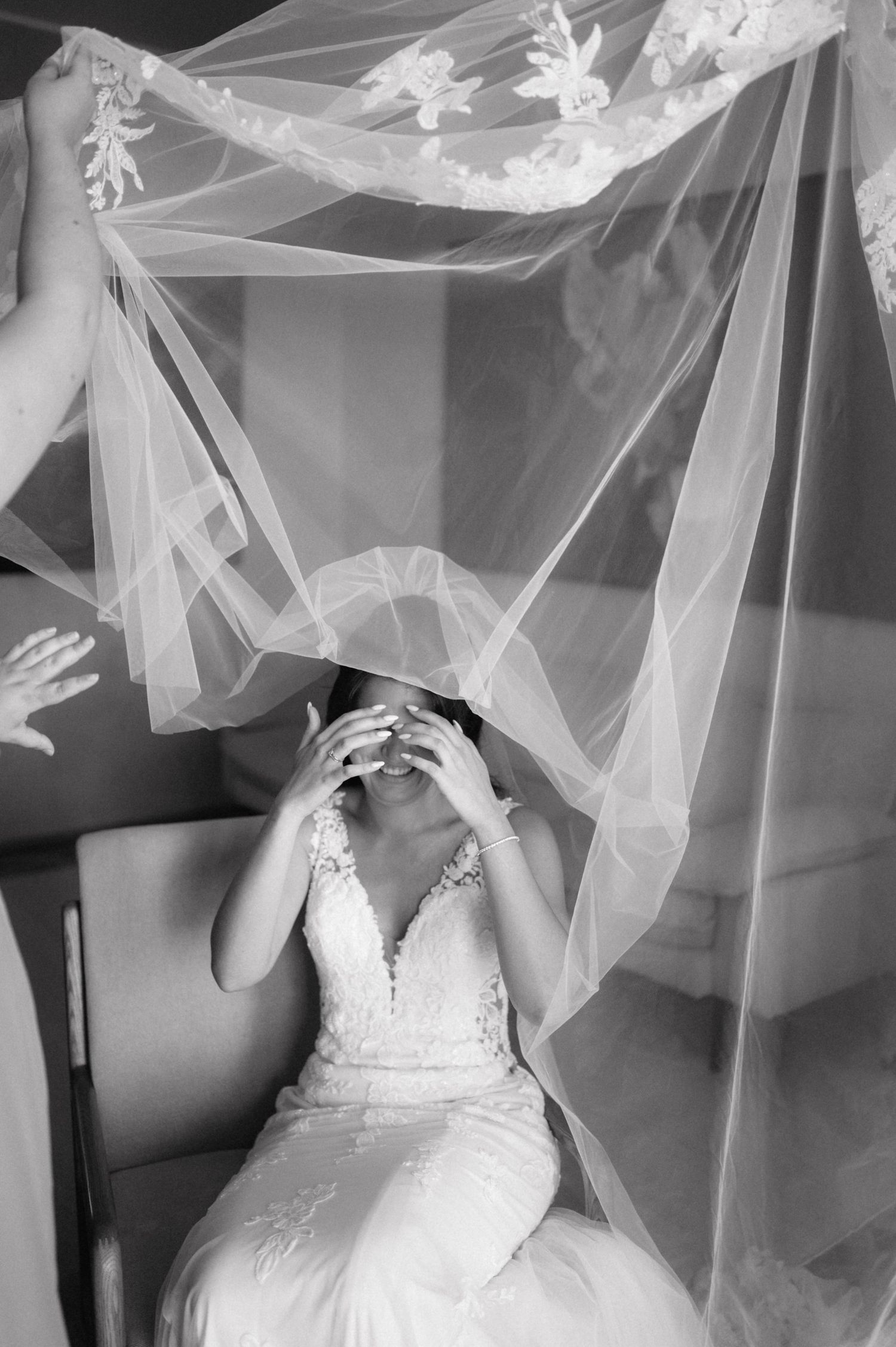 A delicate bridal veil flows gracefully around someone wearing an elegant lace wedding dress with straps against a light background.