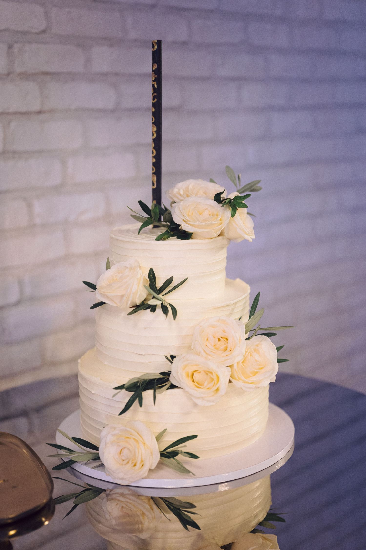 A white wedding cake decorated with fresh pale roses and greenery sits against a white brick wall.