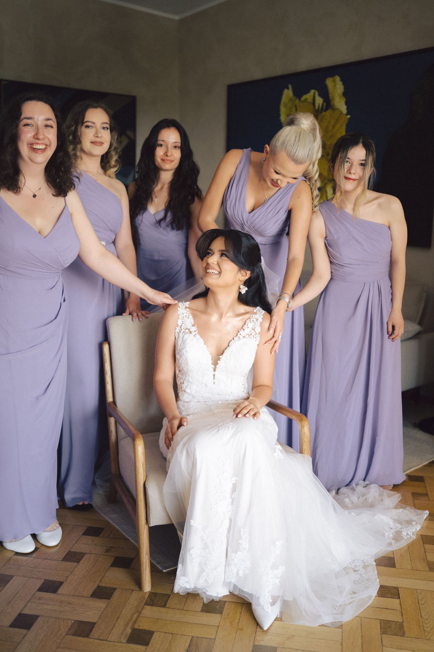 Wedding party wearing lavender bridesmaid dresses gather around bride in white lace gown seated in chair.