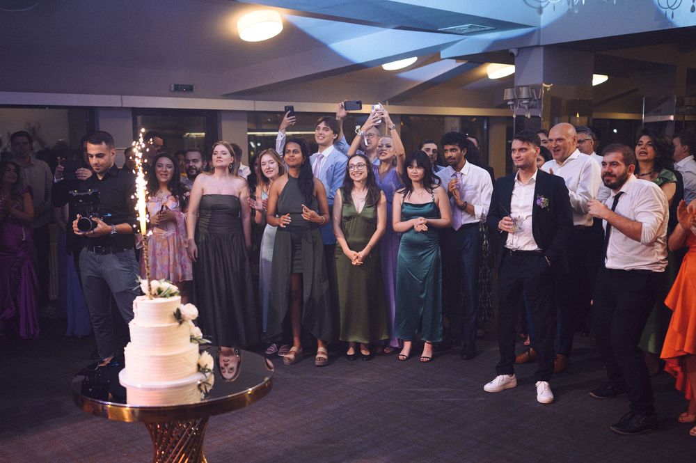 Wedding guests gather in a reception venue watching a sparkler-topped wedding cake being presented.