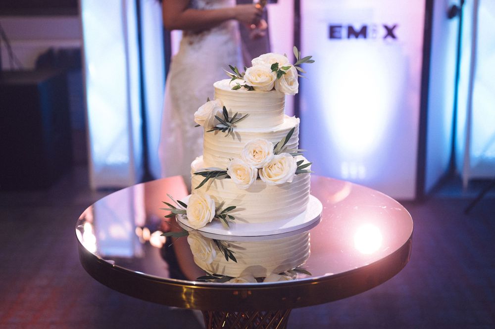 White wedding cake with purple light ambiance and floral accents displayed on a round cake stand in an event venue.