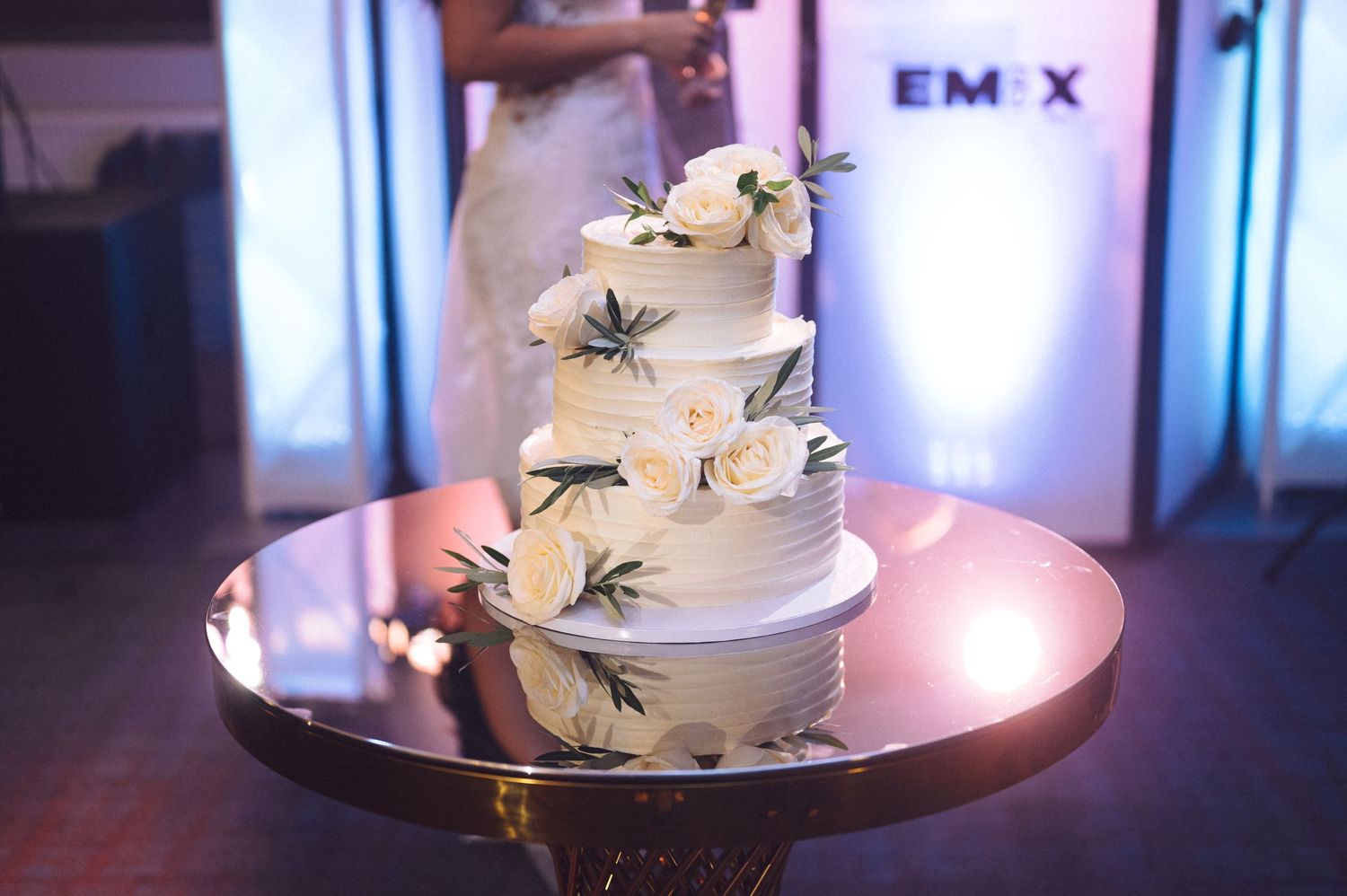 White wedding cake with purple light ambiance and floral accents displayed on a round cake stand in an event venue.