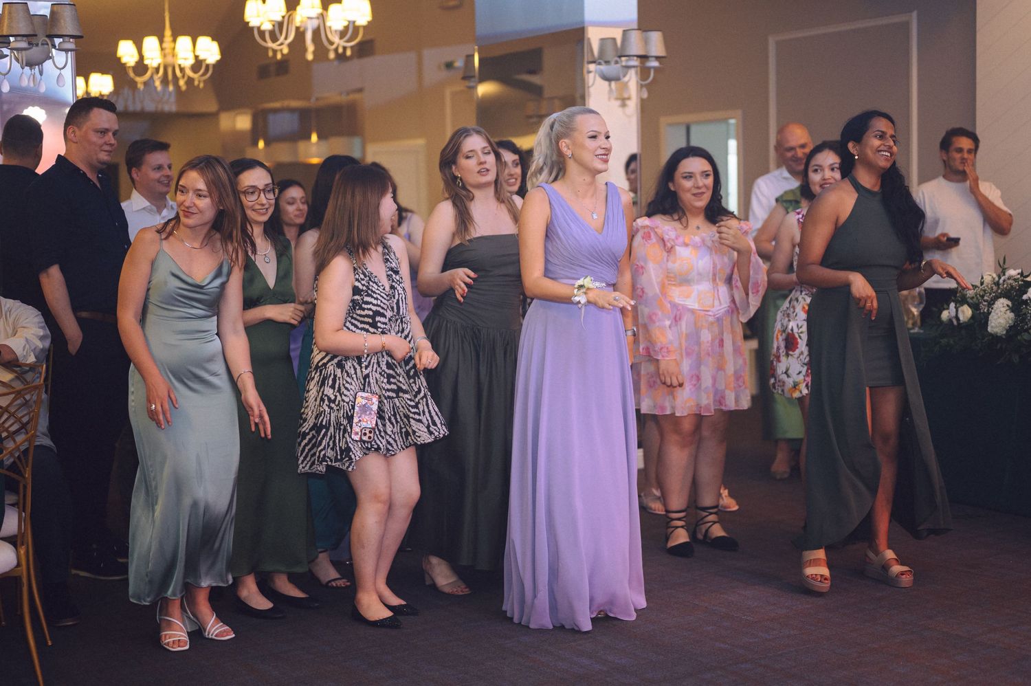 Wedding guests laugh and dance together during a reception in a banquet hall with a formal chandelier overhead.