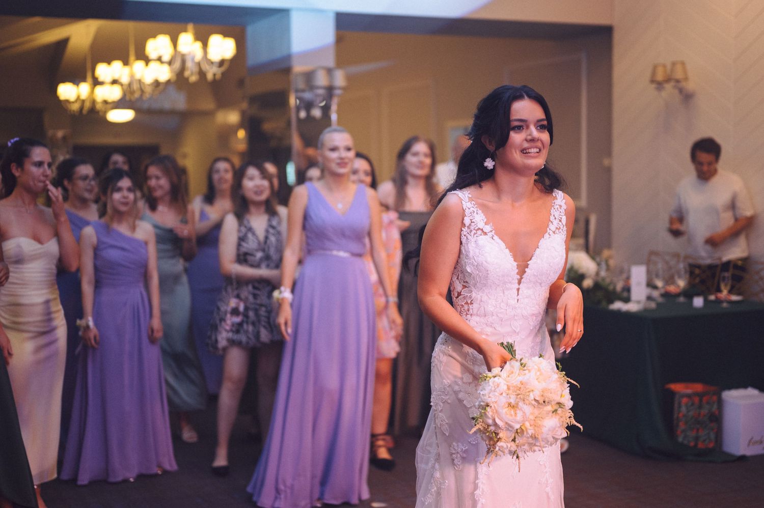 Wedding party gathers during reception as someone prepares to toss bouquet while bridesmaids in purple dresses watch.