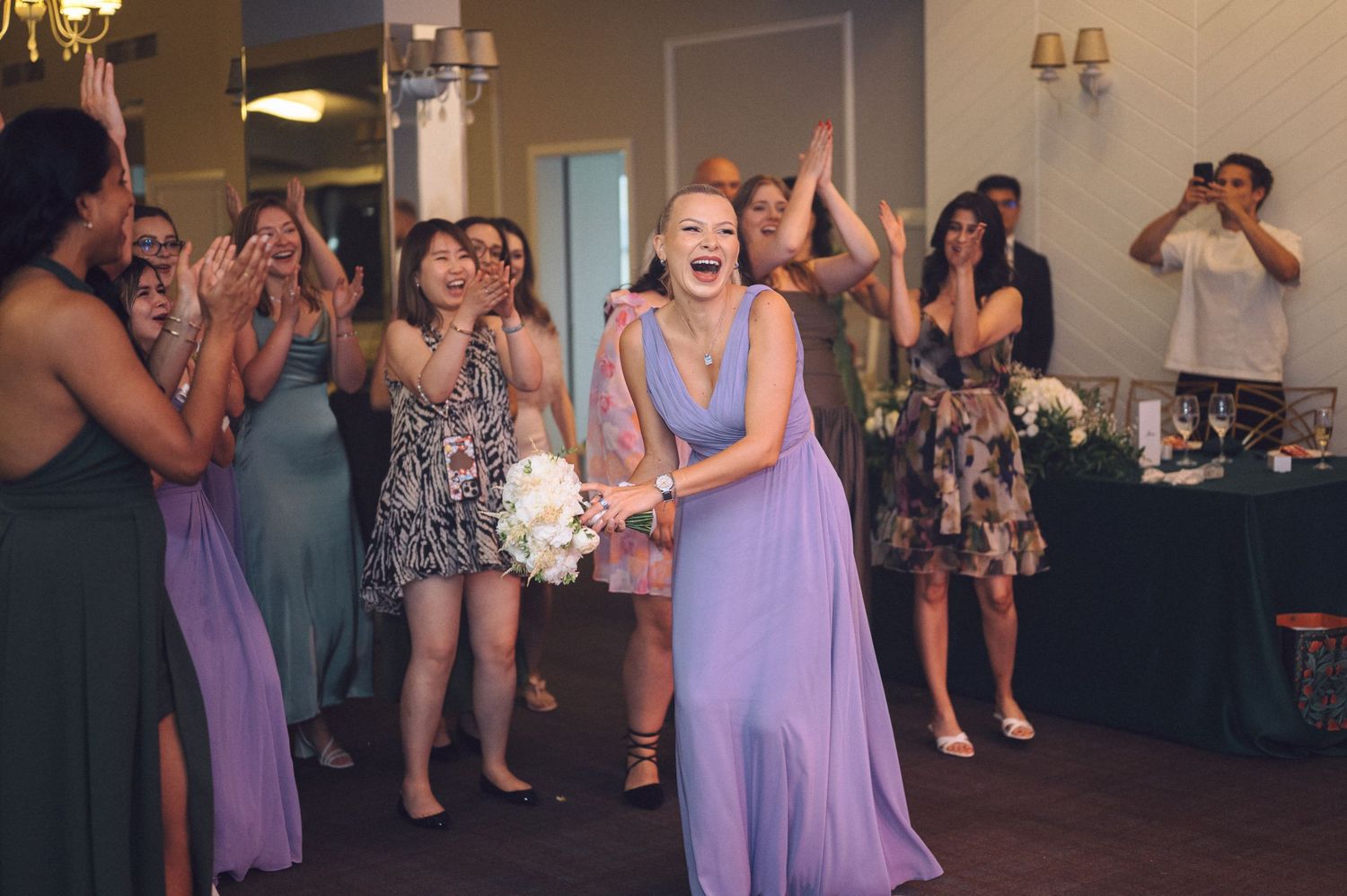 Wedding guests in purple dresses gather excitedly at a reception as someone tosses a bouquet in the air.