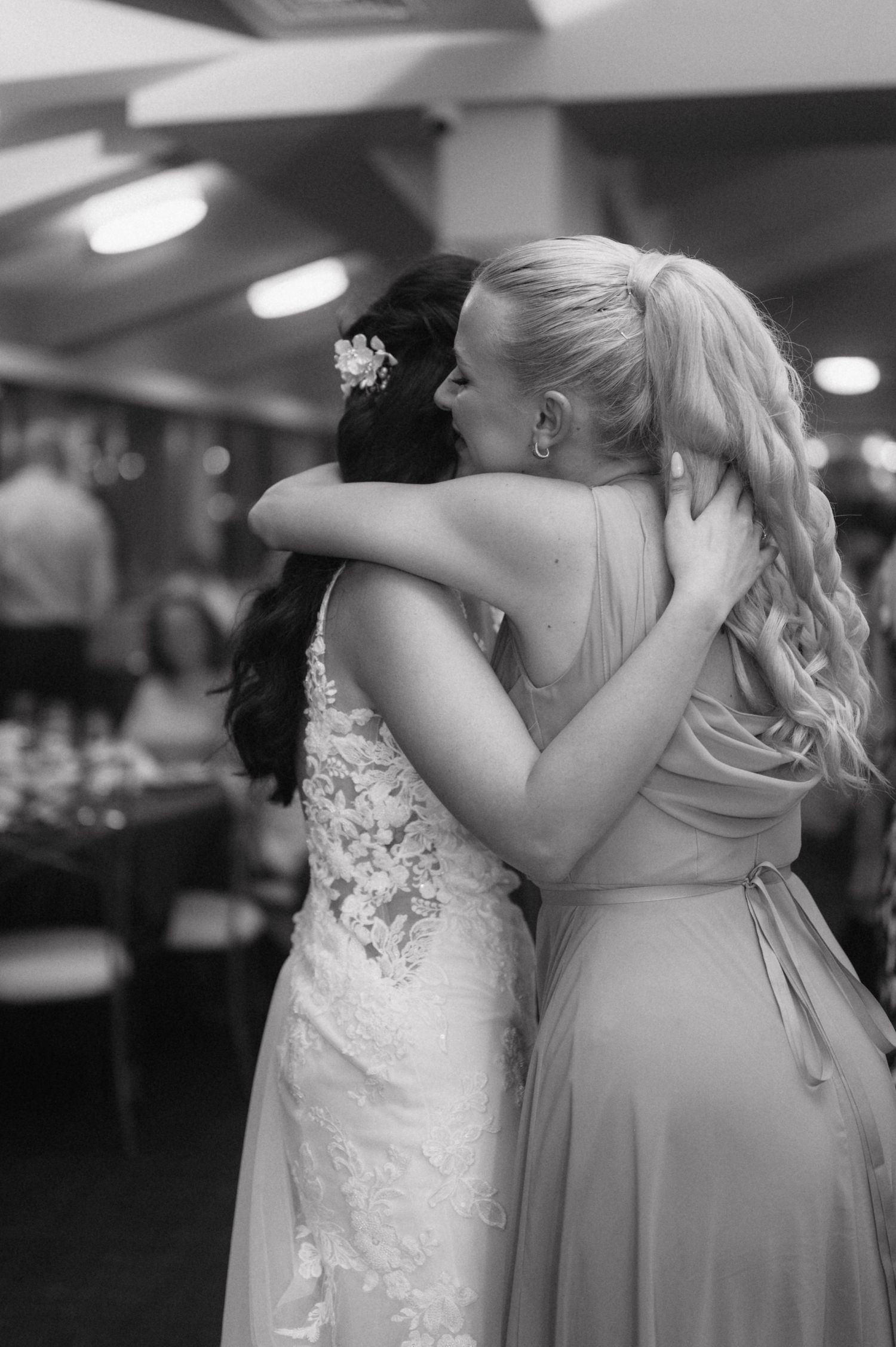 A heartfelt embrace is shared between a bride in a lace gown and a bridesmaid in a light colored dress during a wedding.