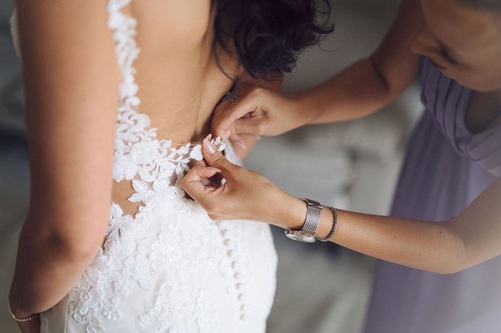 Hands carefully buttoning up delicate white lace wedding dress with intricate details.