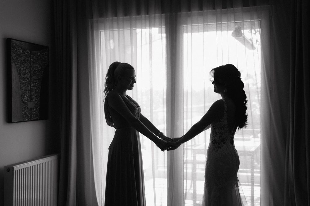 Two silhouetted figures hold hands while standing by a bright window in a black and white artistic photograph.