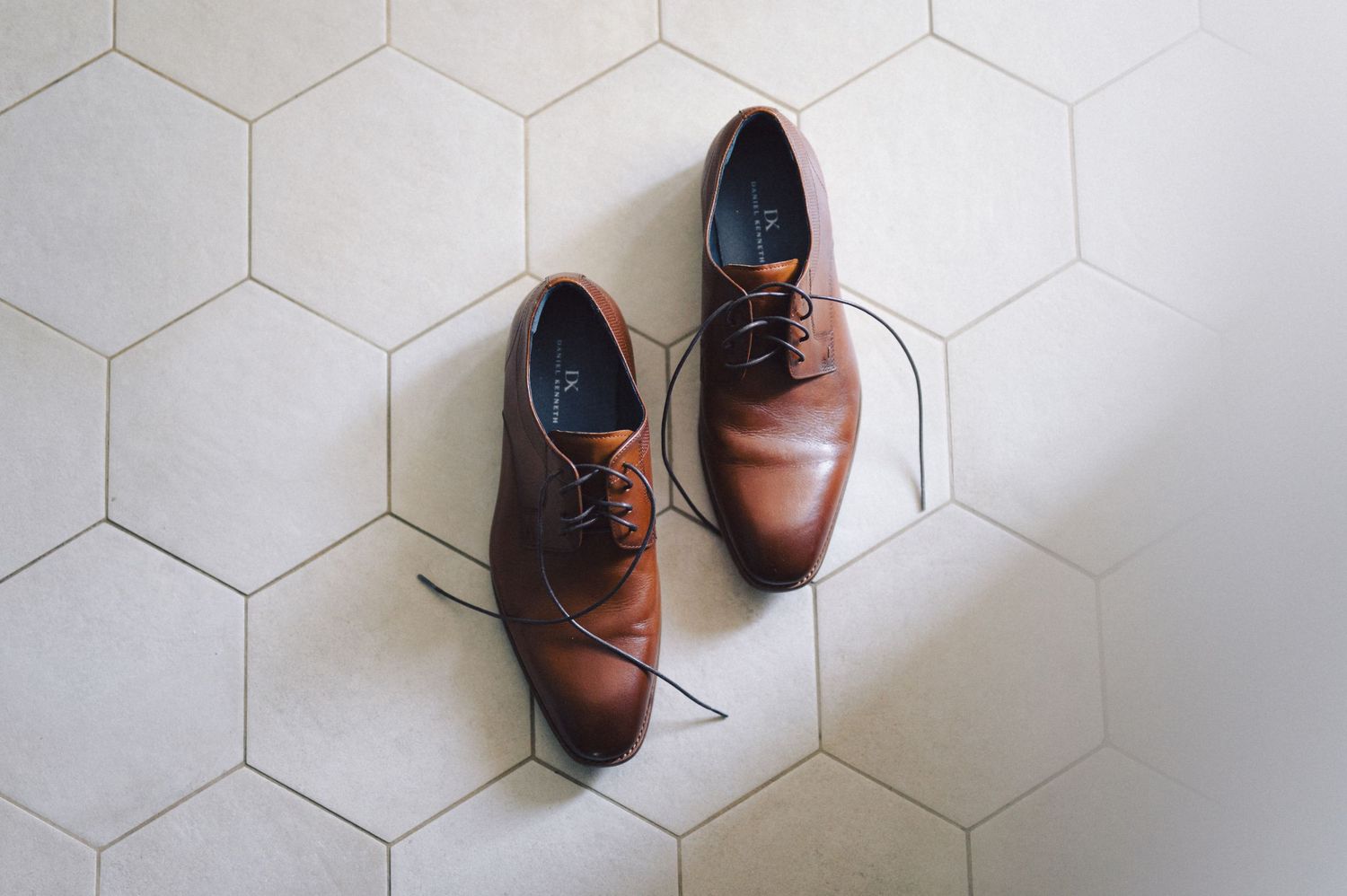 Two brown leather lace-up men's dress shoes arranged in different positions on white hexagonal floor tiles.