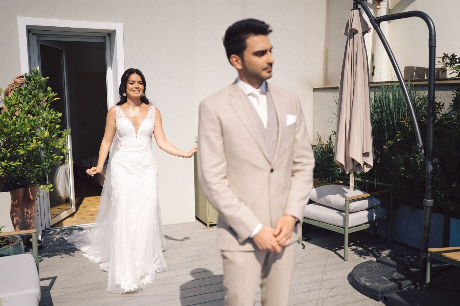 A couple in wedding attire shares a romantic first look moment on an outdoor terrace with potted plants and patio furniture.