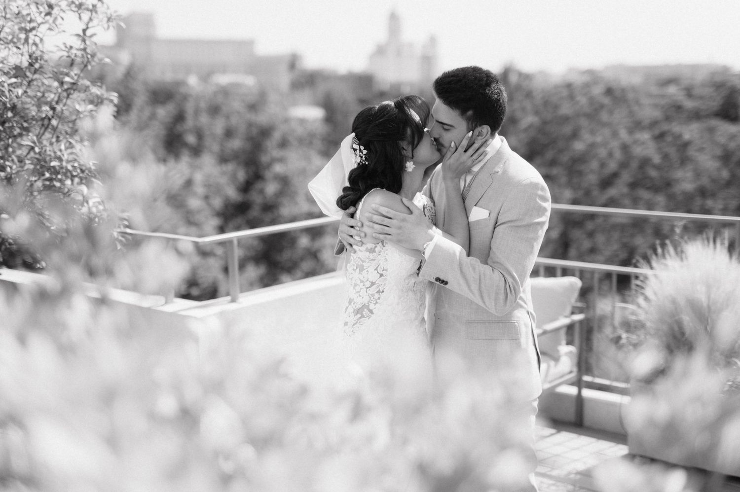 A tender black and white wedding moment captured on a balcony overlooking a cityscape through soft dreamy foliage.