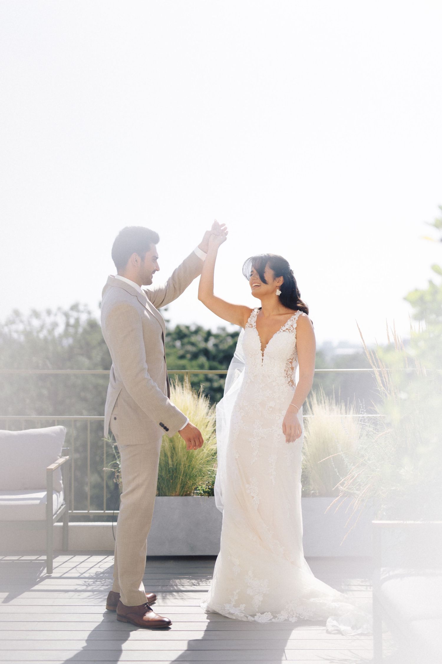 A bride in a white lace gown and groom in a beige suit share a celebratory high five on an outdoor terrace.