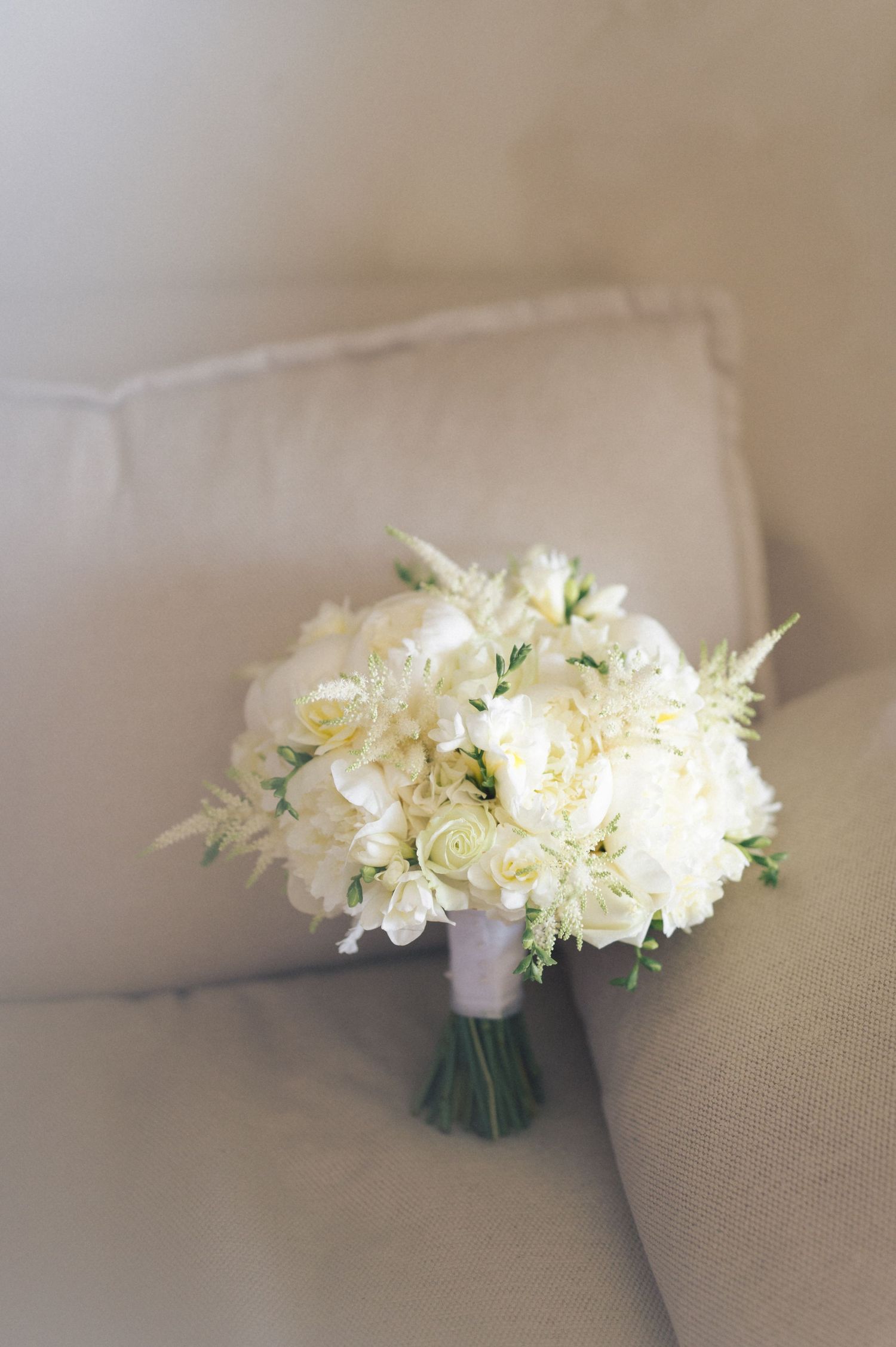 Small white floral boutonniere with delicate blooms and green foliage arranged in a cluster against a beige background.
