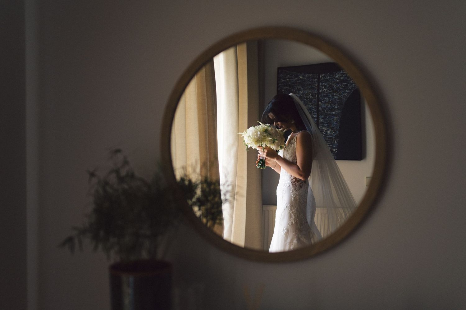 Reflection in a round mirror shows a bride in a white dress holding a bouquet in sunlit room with houseplants.