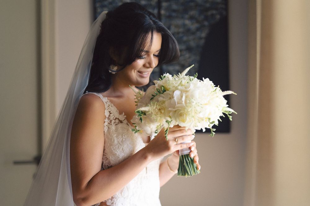 A bride in a white lace gown holds a beautiful white feathered bouquet while smiling in soft window light.