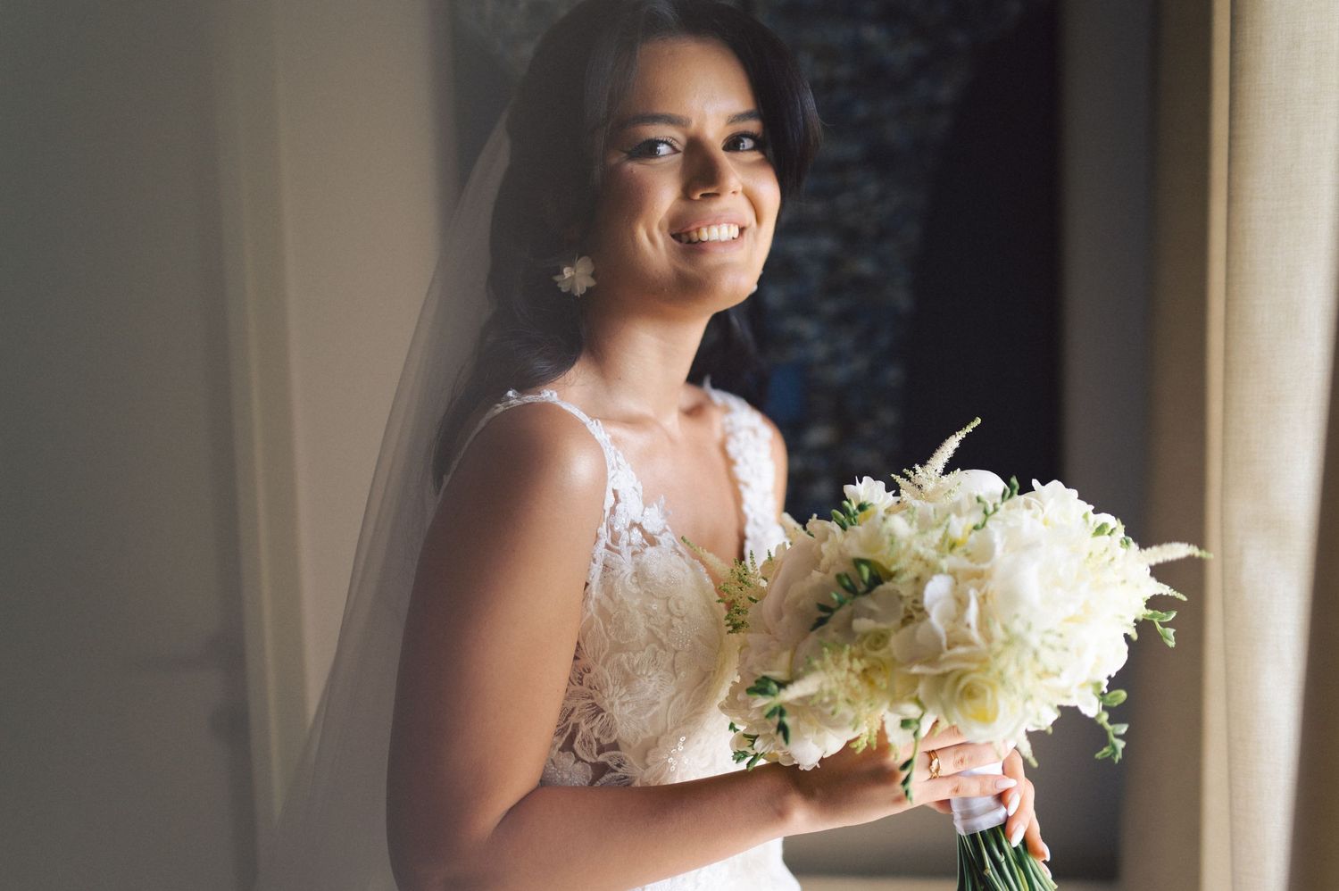 A beautiful bride in a white lace gown holds an elegant bouquet of white flowers while standing near a window.