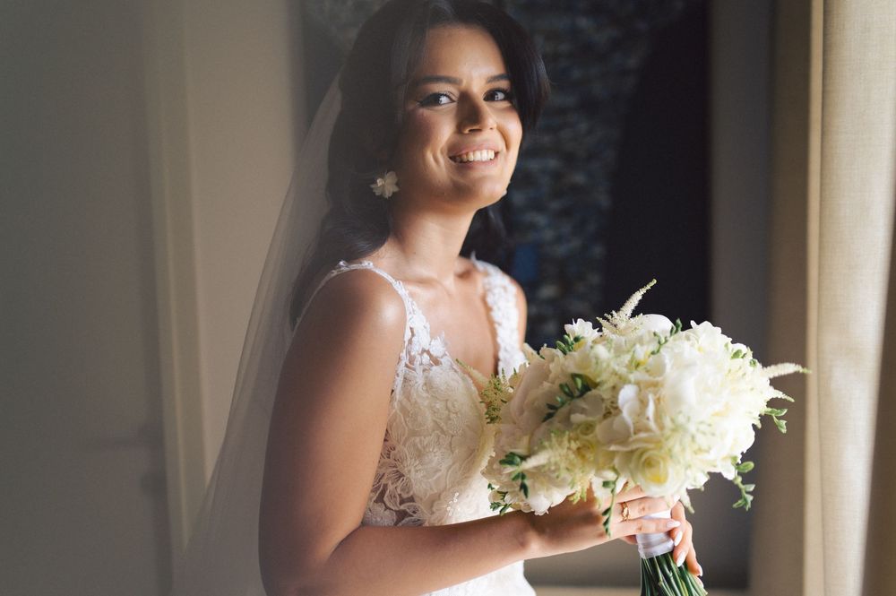 A beautiful bride in a white lace gown holds an elegant bouquet of white flowers while standing near a window.
