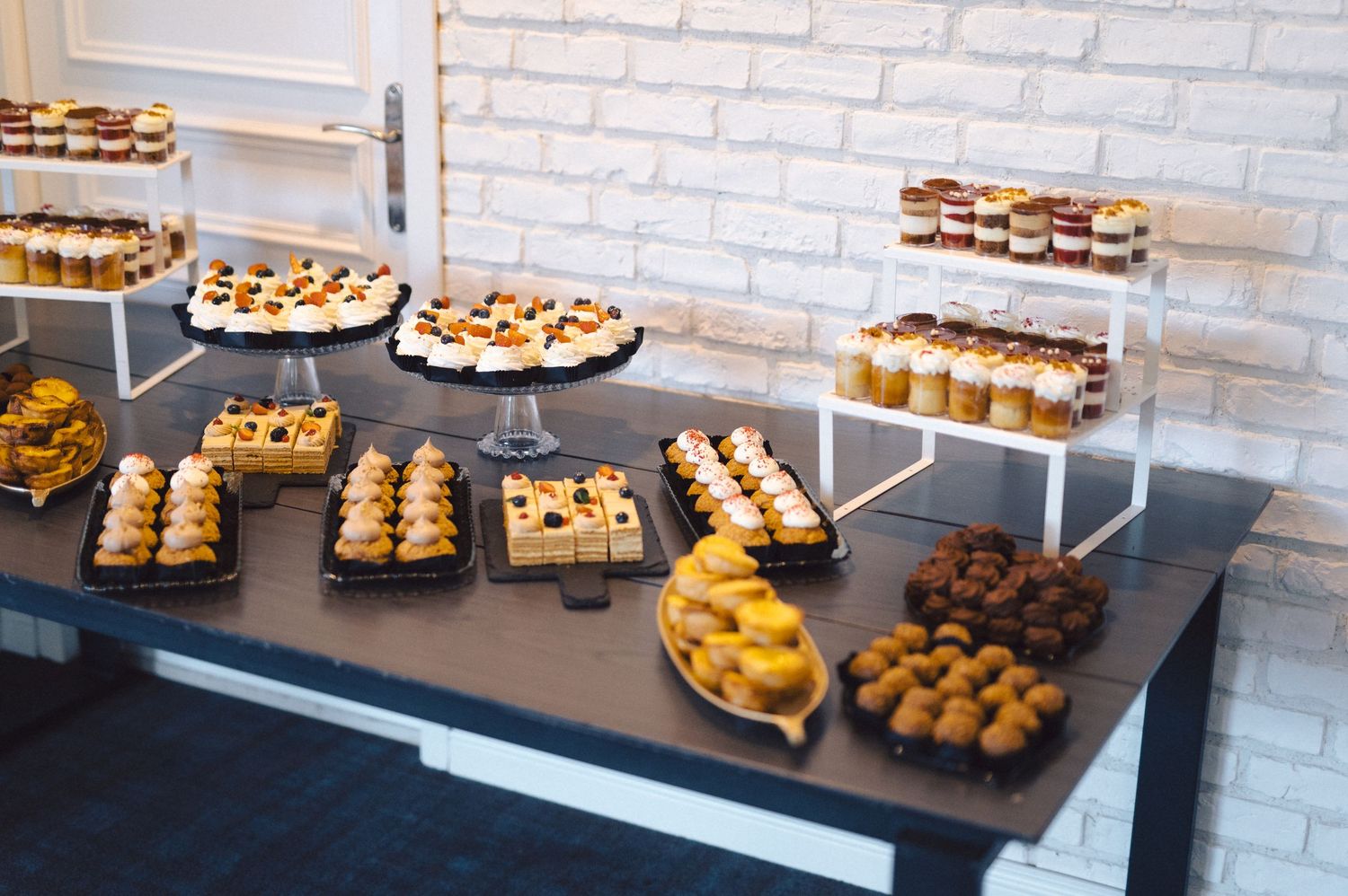 A long buffet table displays an assortment of elegant desserts and pastries against a white brick wall backdrop.