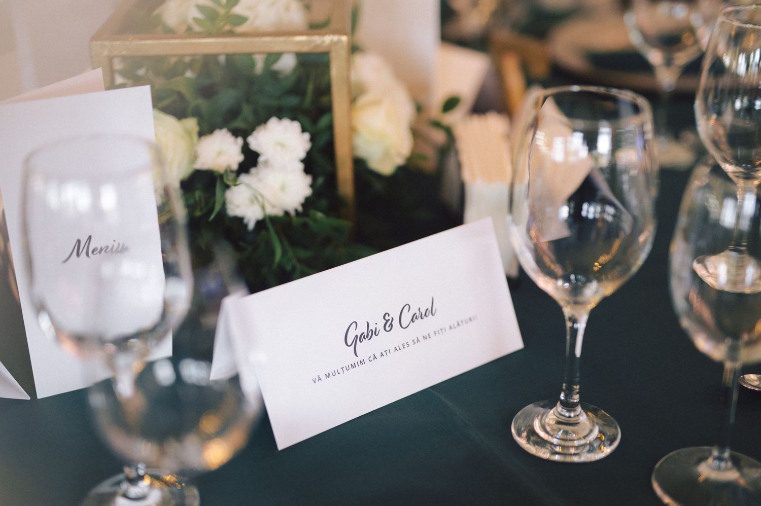 Elegant wedding place setting with menu card, white floral arrangement and wine glasses on dark table.