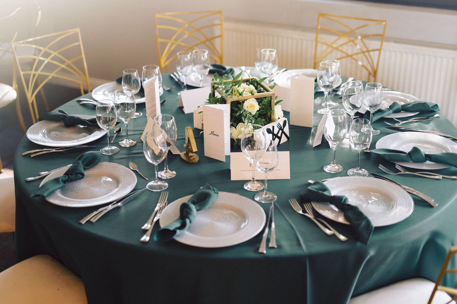 Elegant wedding table setting with dark green tablecloth, white plates, gold chairs and floral centerpiece.