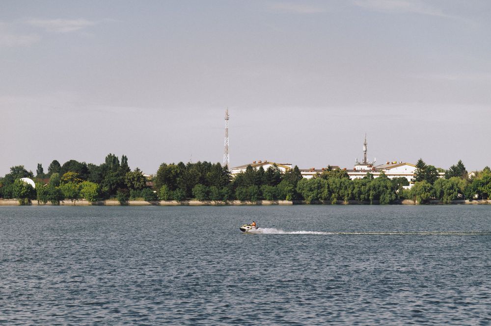 A speedboat cruises across a calm lake with a treeline and buildings visible along the distant shoreline.