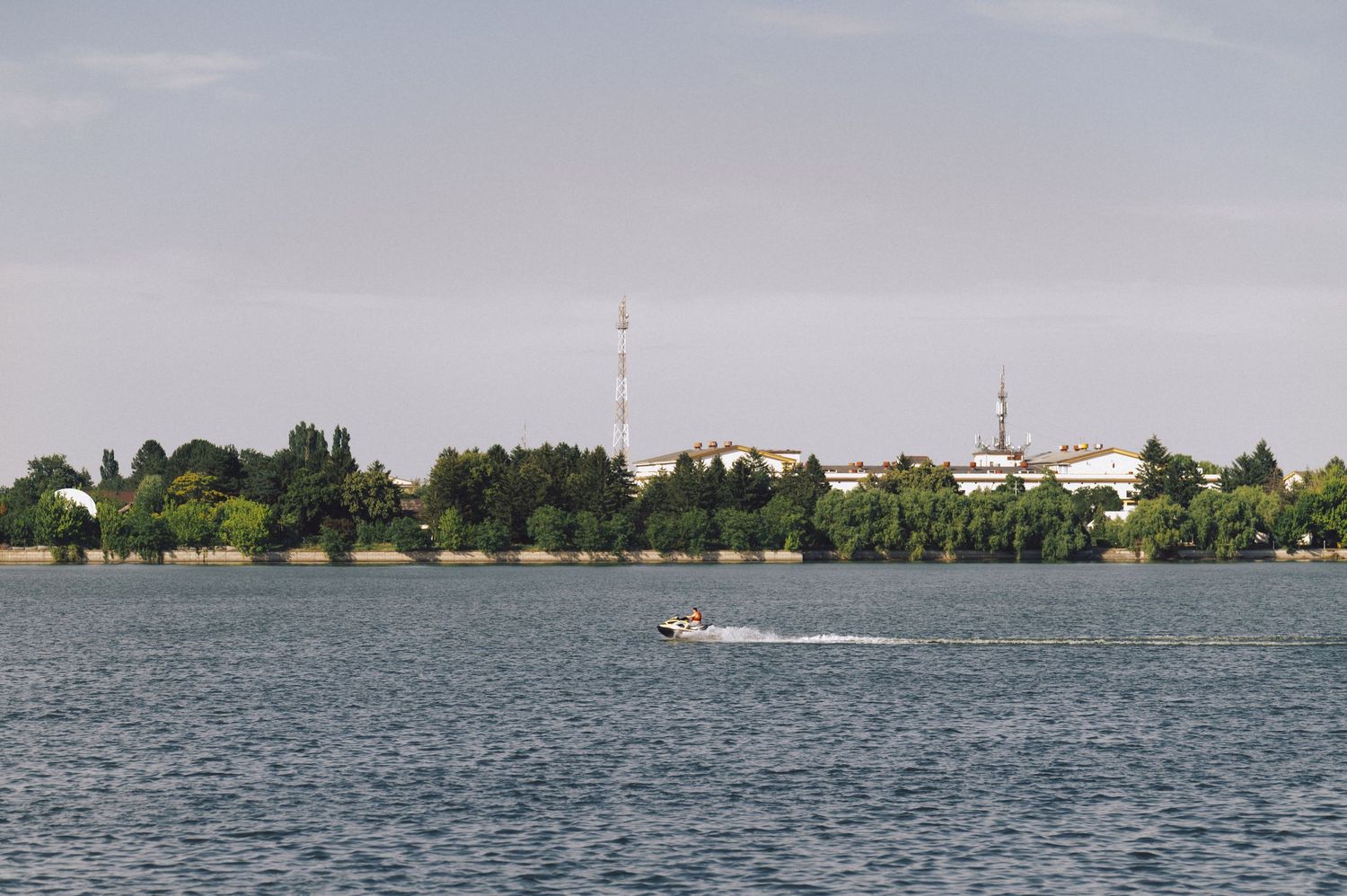 A speedboat cruises across a calm lake with a treeline and buildings visible along the distant shoreline.