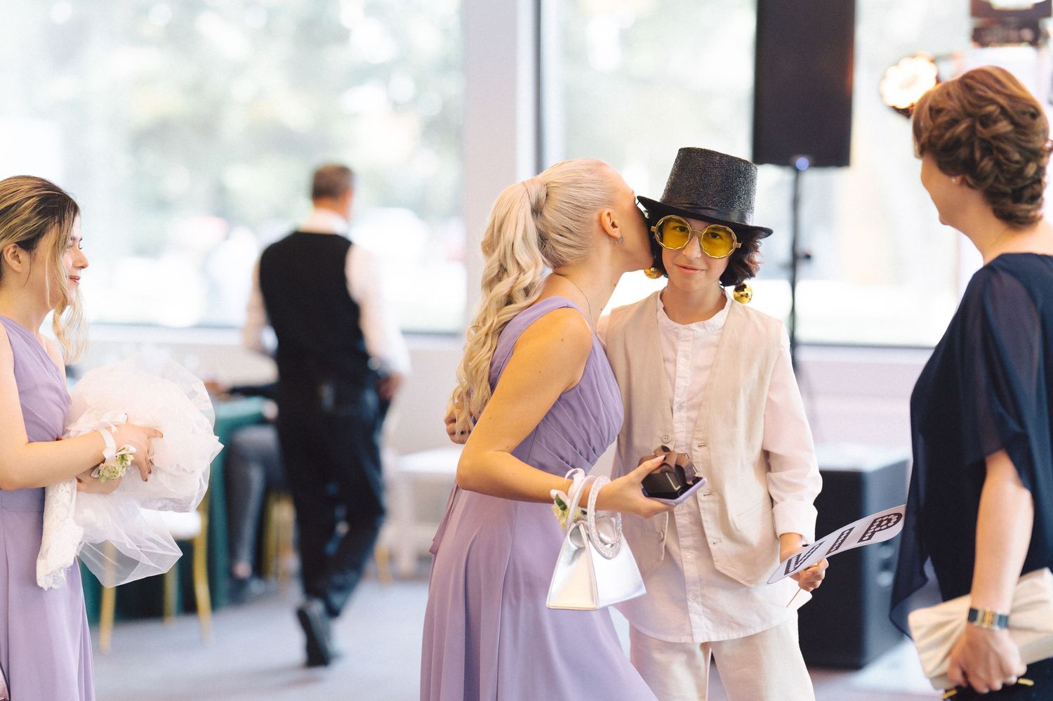 Wedding party gathers in a light-filled venue with people wearing soft lavender and white attire.